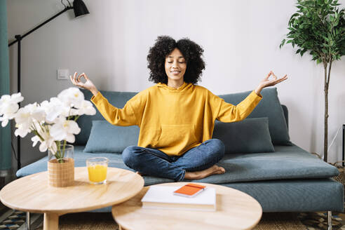 Young woman meditating while sitting on sofa at home - JCZF00636