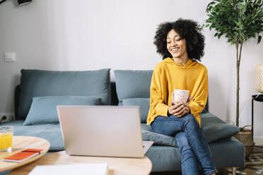 Smiling woman with mug looking at laptop while sitting at home - JCZF00637