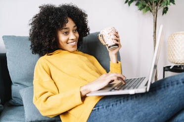 Young woman using laptop while holding mug at home - JCZF00638