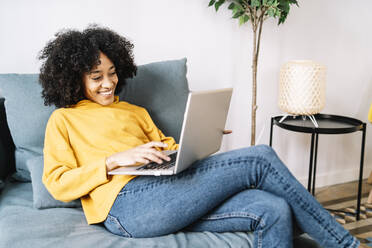 Lächelnde junge Frau, die einen Laptop benutzt, während sie zu Hause auf dem Sofa sitzt - JCZF00639