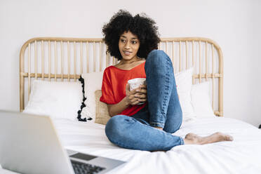 Woman holding mug looking at laptop while sitting on bed at home - JCZF00652