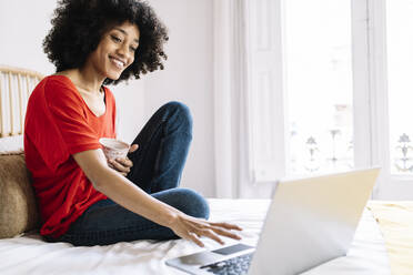 Junge Frau mit Tasse, die einen Laptop benutzt, während sie zu Hause auf dem Bett sitzt - JCZF00653