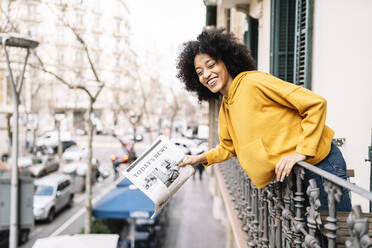 Cheerful young woman with newspaper leaning on railing in balcony - JCZF00666