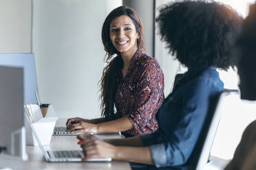 Lächelnde weibliche Fachkraft, die einen Kollegen bei der Arbeit am Laptop im Büro ansieht - JSRF01492