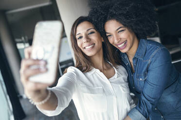 Fröhliche Geschäftsfrau, die eine Kollegin umarmt, während sie ein Selfie mit ihrem Mobiltelefon im Büro macht - JSRF01502