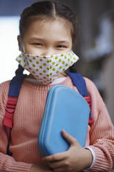 Mädchen mit Gesichtsschutzmaske, das während einer Pandemie ein Mäppchen hält - AZF00225