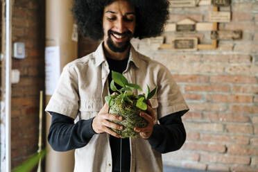 Lächelnder Afro-Mann mit Blick auf eine Kokedama-Zimmerpflanze - AFVF08495