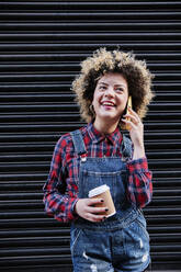 Happy woman holding disposable coffee cup while talking on smart phone in front of shutter - ASGF00159