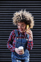 Curly haired woman with disposable coffee cup laughing while talking on mobile phone - ASGF00160