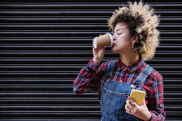 Frau in Latzhose trinkt Kaffee, während sie ihr Handy an den Auslöser hält - ASGF00161