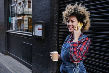 Smiling woman with disposable coffee cup looking away while talking on mobile phone in front of shutter - ASGF00165