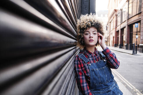 Young woman with hand in hair leaning on corrugated iron - ASGF00172