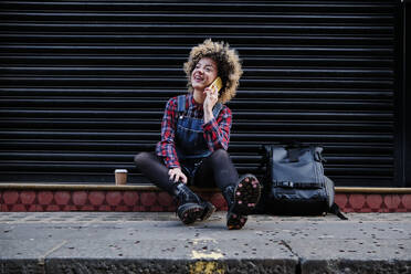 Smiling curly haired woman talking on mobile phone while sitting on footpath - ASGF00176