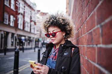 Junge Frau mit Sonnenbrille, die ihr Smartphone benutzt, während sie sich im Urlaub in Chinatown an die Wand lehnt - ASGF00188