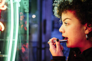 Young woman applying lipstick while looking at neon lights - ASGF00193