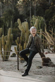 Älterer Mann, der wegschaut, während er sich auf ein Geländer vor einem Kaktusgarten stützt - GMLF01086