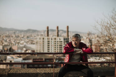 Älterer Mann mit Jacke, der auf einer Stützmauer sitzt, während die Stadt im Hintergrund zu sehen ist - GMLF01093