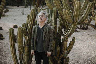 Mature man with eyes closed standing amidst cactus plants in garden - GMLF01096