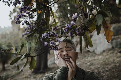 Älterer Mann, der sein Gesicht berührt, während er unter einem blühenden Baum im Garten steht - GMLF01100