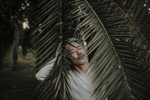 Älterer Mann mit geschlossenen Augen inmitten von Palmenblättern - GMLF01110