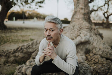 Gray haired man sitting on tree roots in garden - GMLF01117