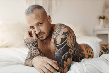 Serious man with tattoo lying on bed at home - GMLF01125