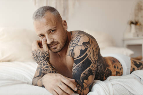 Ernster Mann mit Tattoo auf dem Bett liegend zu Hause - GMLF01125