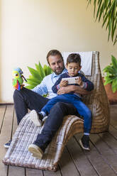 Smiling father looking at son playing with mobile phone on lounge chair - DLTSF01715