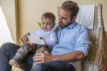 Father sharing mobile phone with cute son sitting on lap at home - DLTSF01717