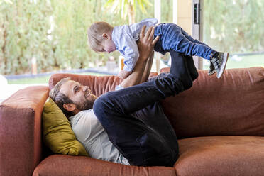 Smiling father lifting son while lying on sofa at living room - DLTSF01719