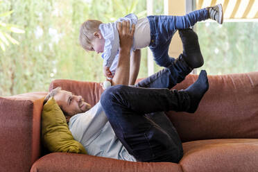 Smiling father lifting playful son on sofa at home - DLTSF01720