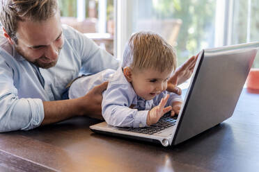 Smiling father and son waving during video call through laptop at home - DLTSF01722