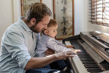 Father teaching piano to son sitting on lap at home - DLTSF01732