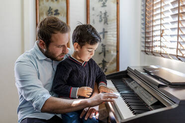 Boy learning piano sitting on father's lap at home - DLTSF01733