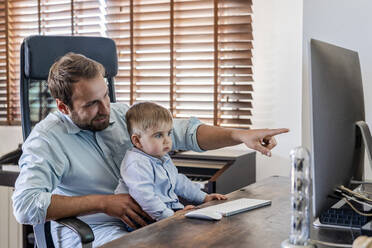 Lächelnder Vater, der auf einen Computer zeigt, während sein Sohn zu Hause auf dem Schoß sitzt - DLTSF01736