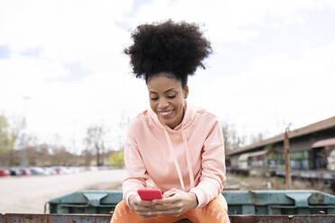 Lächelnde Frau, die ein Smartphone benutzt, während sie im Freien sitzt - JCCMF01750