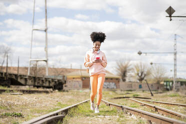 Smiling woman using smart phone while walking on railway track during sunny day - JCCMF01766