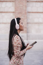 Woman with eyes closed holding smart phone while standing by wall - EBBF03183