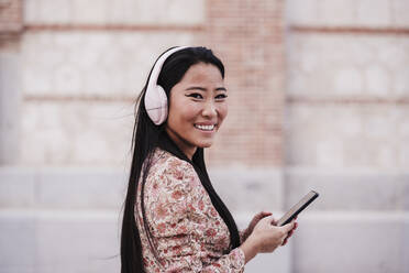 Smiling woman with headphones holding smart phone by wall - EBBF03184