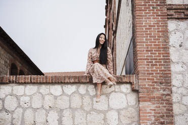 Carefree woman sitting on retaining wall in city - EBBF03206