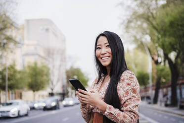 Woman looking away while holding smart phone in city - EBBF03216