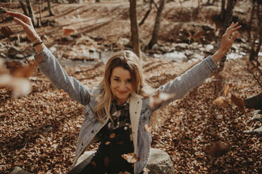 Smiling woman playing with autumn leaves while sitting on rock in forest - GMLF01137