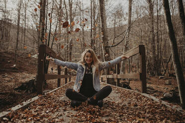 Happy woman playing with autumn leaves while sitting on wooden bridge in forest - GMLF01144