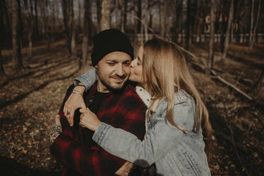 Woman in denim jacket kissing hipster boyfriend in forest during autumn - GMLF01150