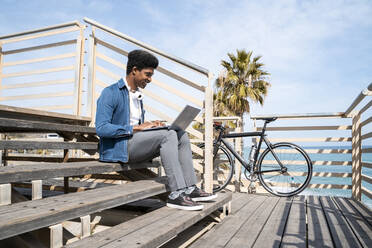 Smiling man using laptop while sitting on steps at promenade by bicycle during  sunny day - AFVF08504