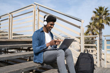 Glücklicher Mann gestikuliert während eines Videogesprächs am Laptop, während er an einem sonnigen Tag auf der Promenade sitzt - AFVF08505