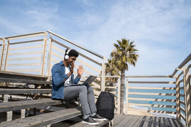 Mann führt Videogespräch auf Laptop, während er an einem sonnigen Tag auf der Promenade sitzt - AFVF08506
