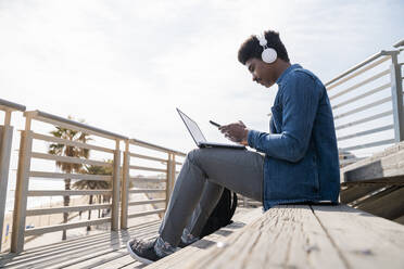 Afro-Mann, der ein Smartphone über einem Laptop benutzt, während er auf den Stufen einer Promenade sitzt - AFVF08510