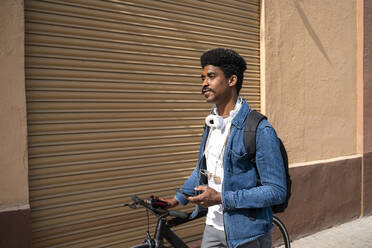 Afro-Mann mit Fahrrad hält Smartphone an der Wand während eines sonnigen Tages - AFVF08515