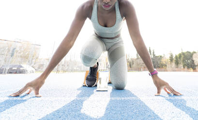 From below of African American female runner in starting blocks sitting in crouch position while getting ready for sprint at stadium during workout - ADSF22518